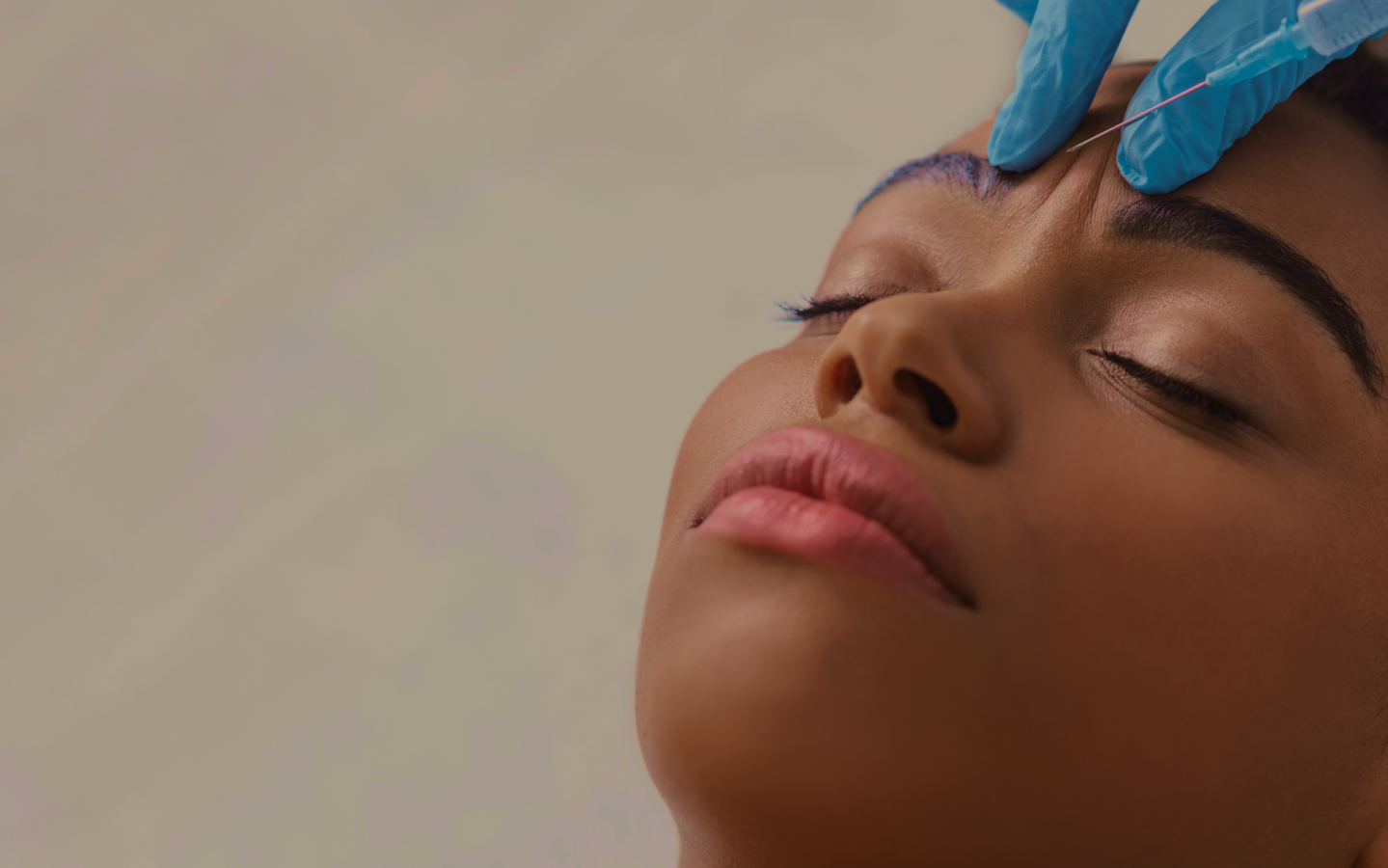 Close-up of a woman's face during a cosmetic procedure; gloved hand with needle near her forehead.