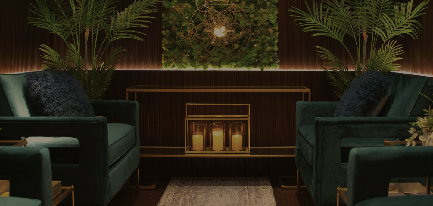Dim lounge with green velvet chairs, gold accents, candles in a glass holder, and plants.