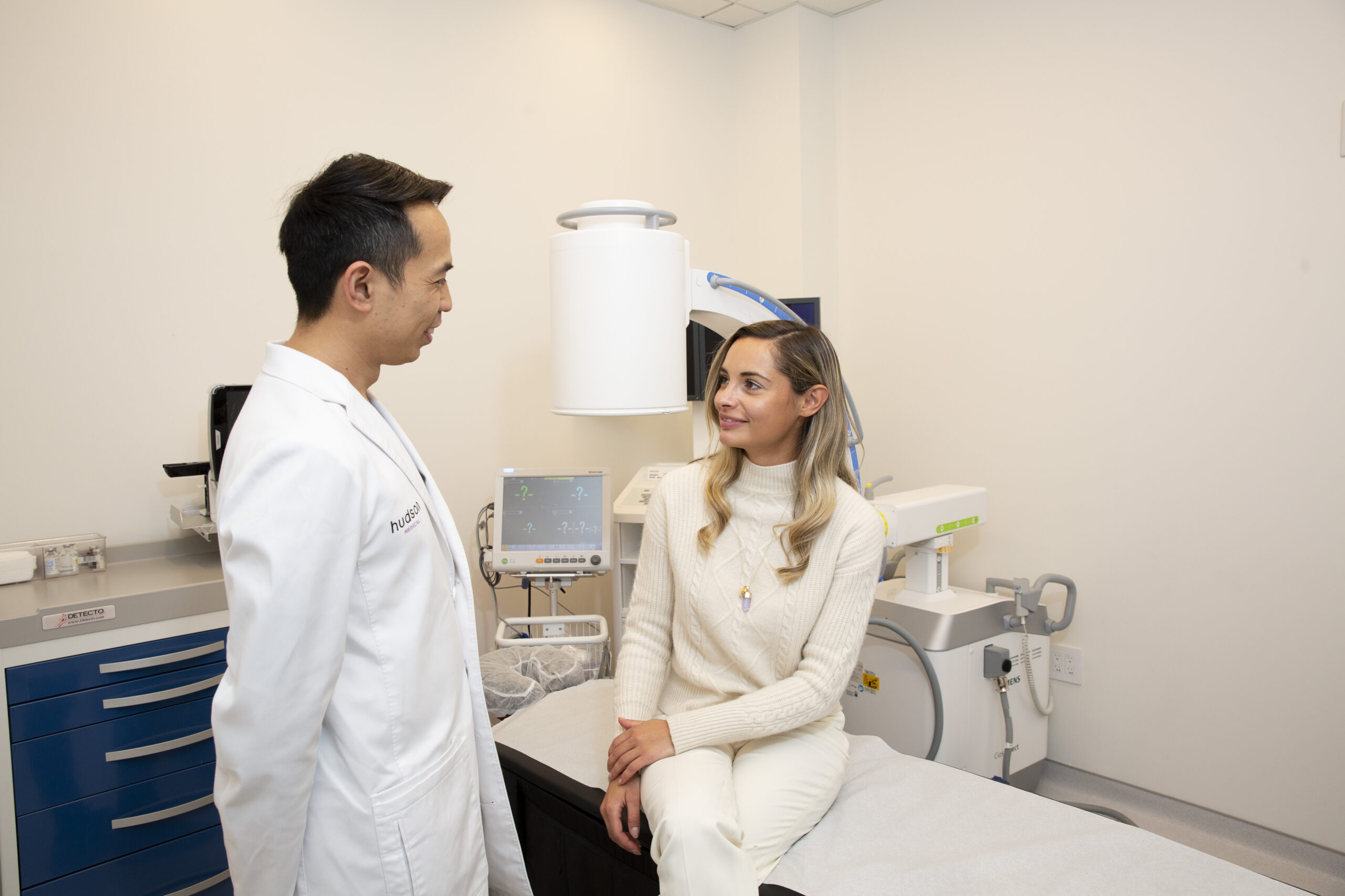 Doctor in white coat and patient sit near radiology imaging equipment in clinic.