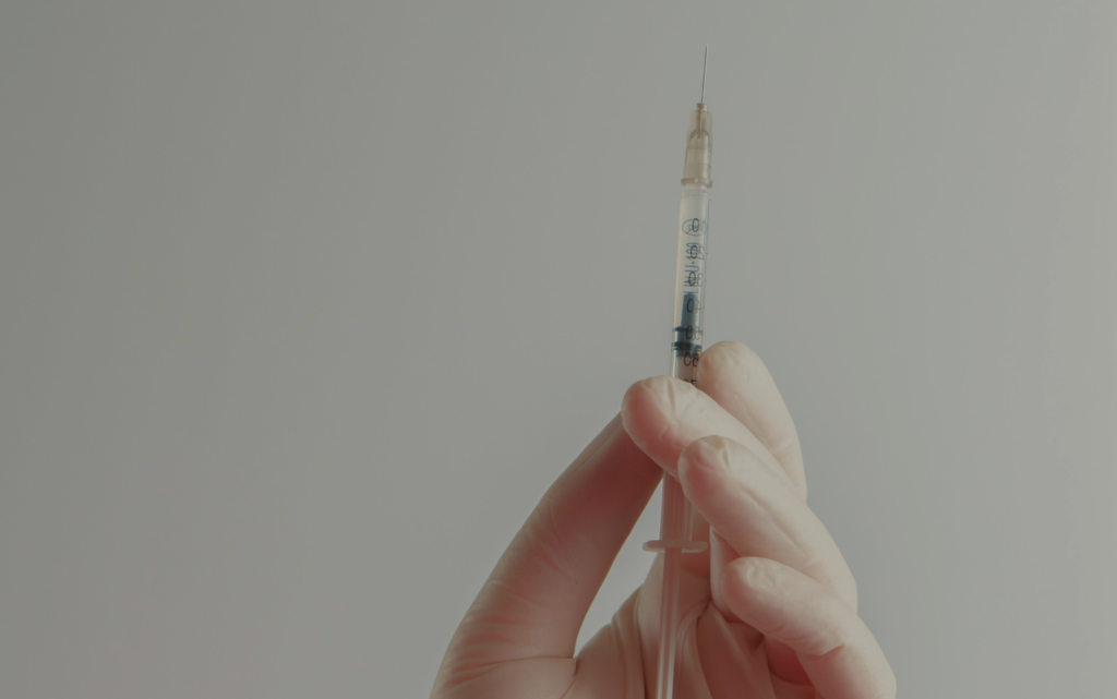 Gloved hand holding a syringe with needle upright against a light background.