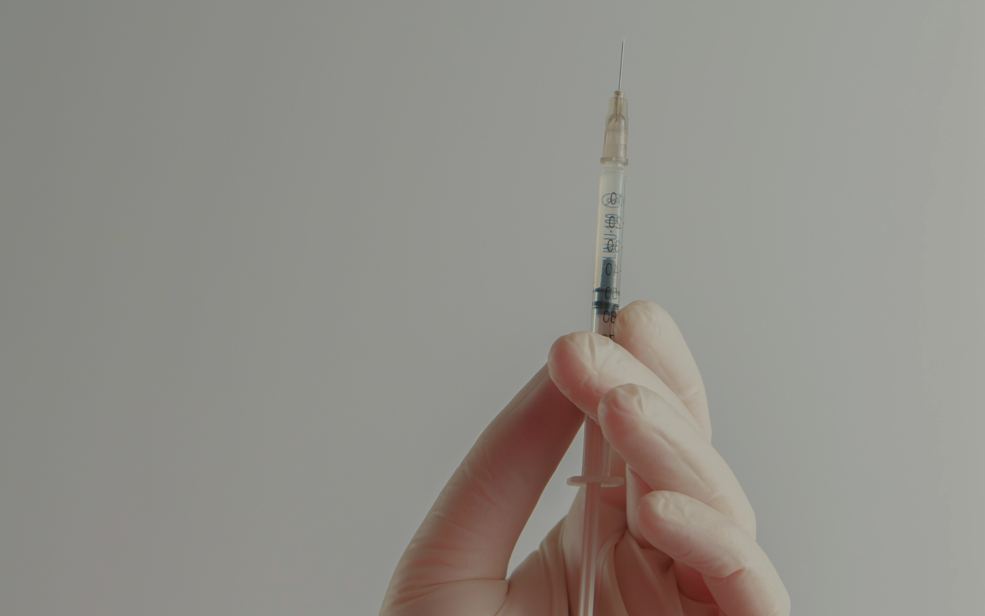 Gloved hand holding a syringe with needle upright against a light background.