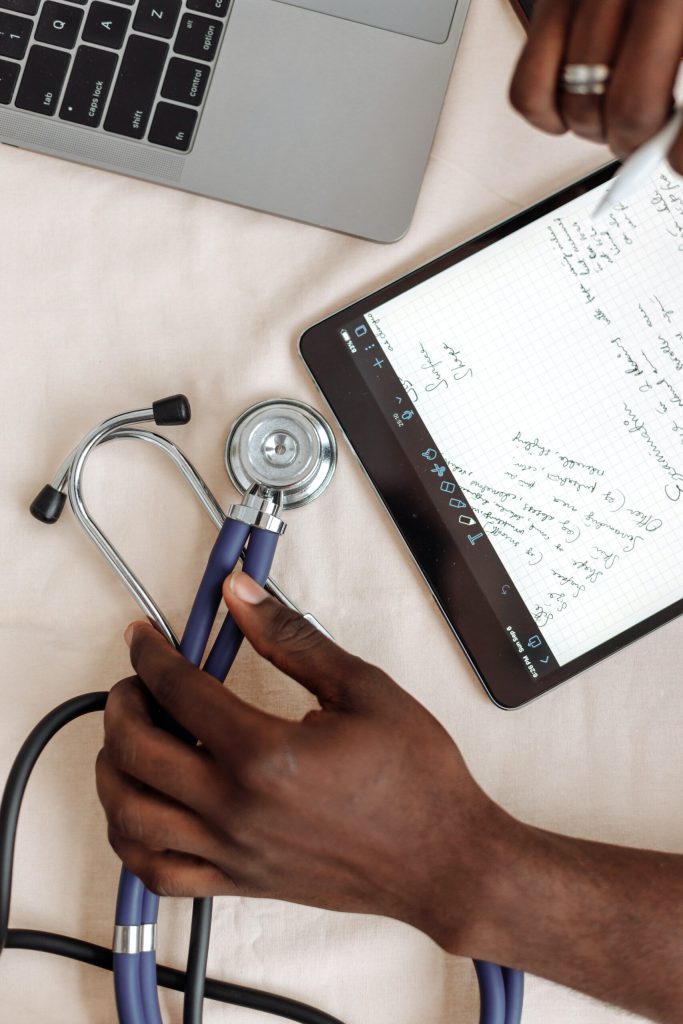 Hands hold a stethoscope beside a tablet with handwritten notes and laptop on a light surface.