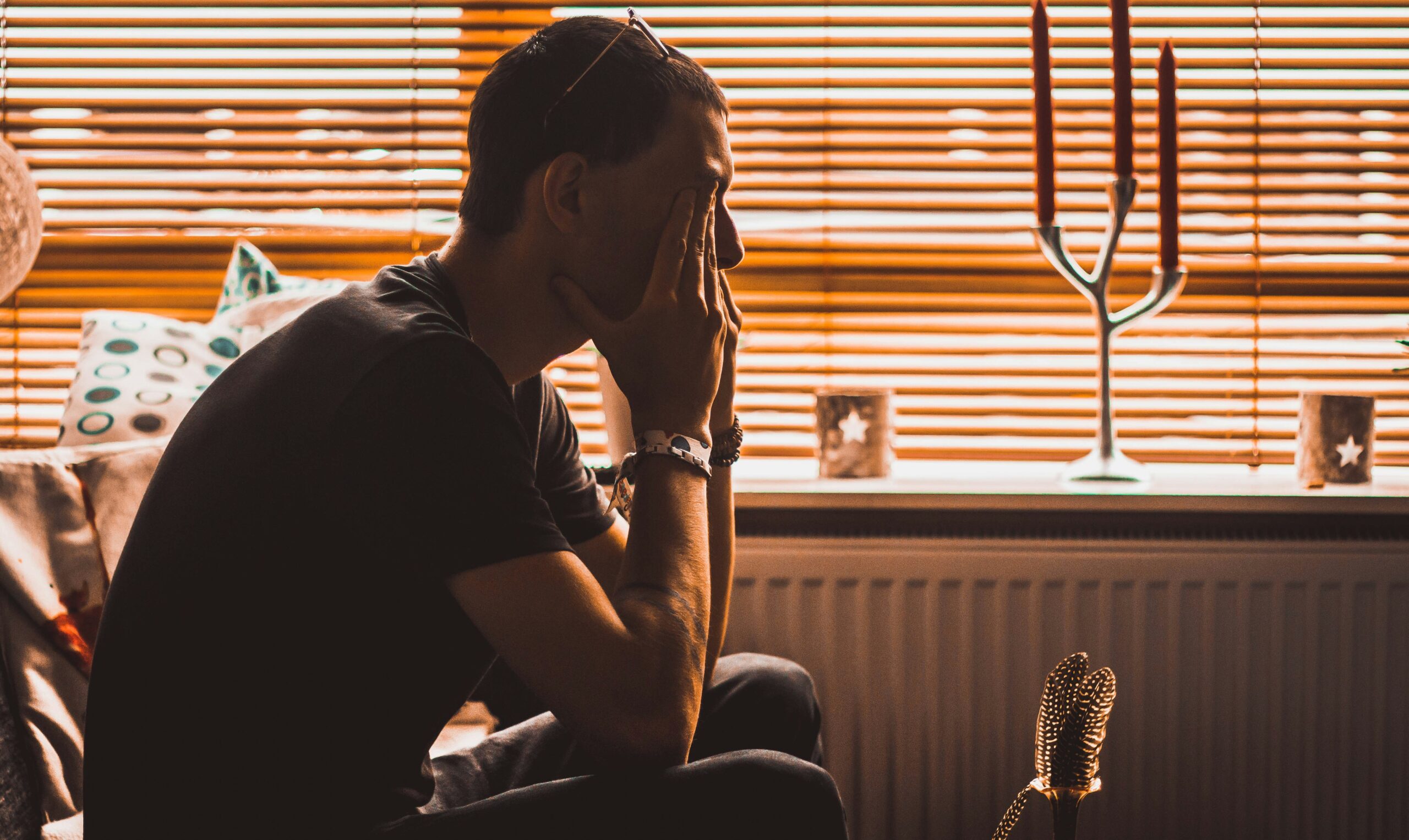 Man sits indoors with face in hands, sunglasses on head, in front of orange blinds.