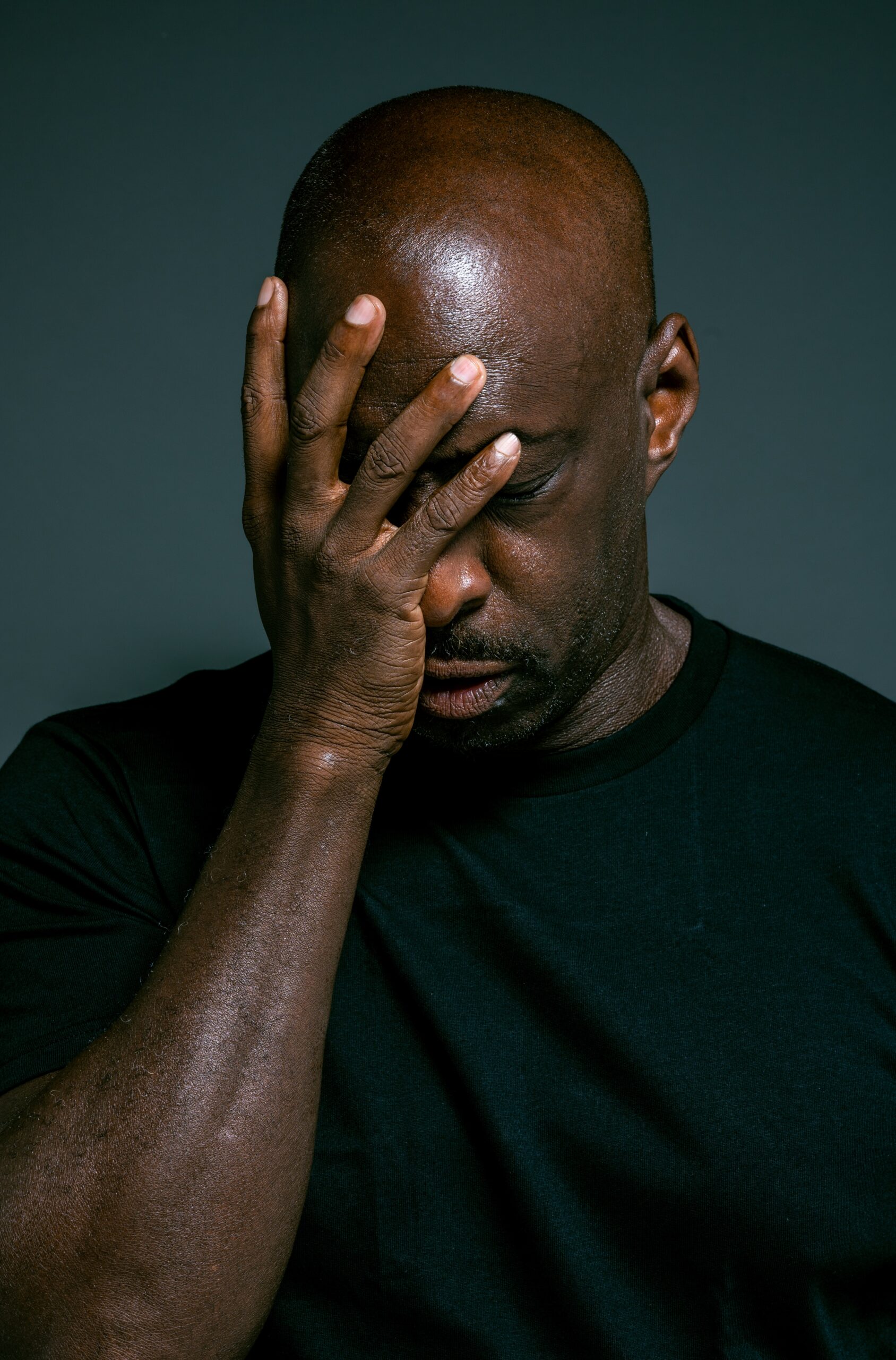 Man with shaved head covers face with hand, looking downward against dark background.