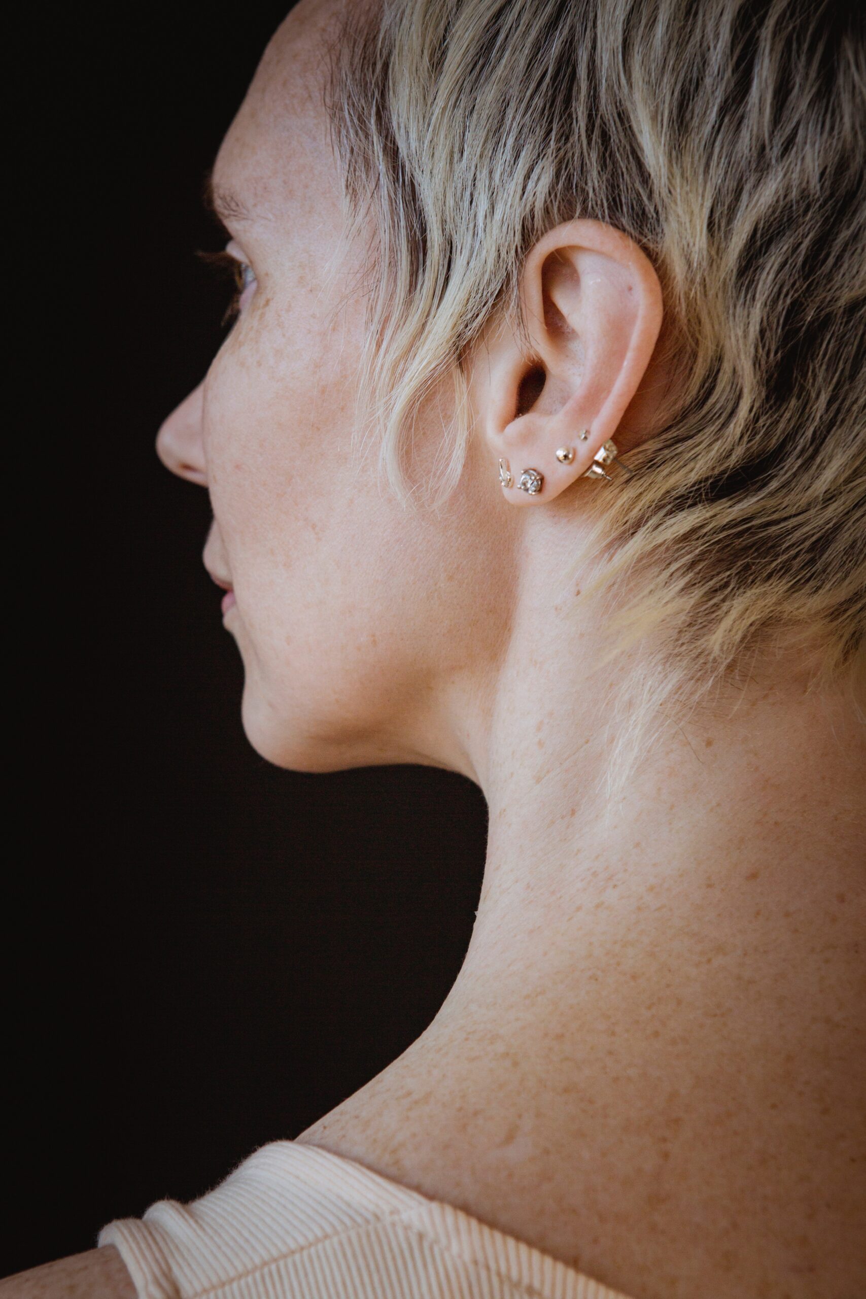Side view of a woman's ear with multiple small earrings and short blonde hair.