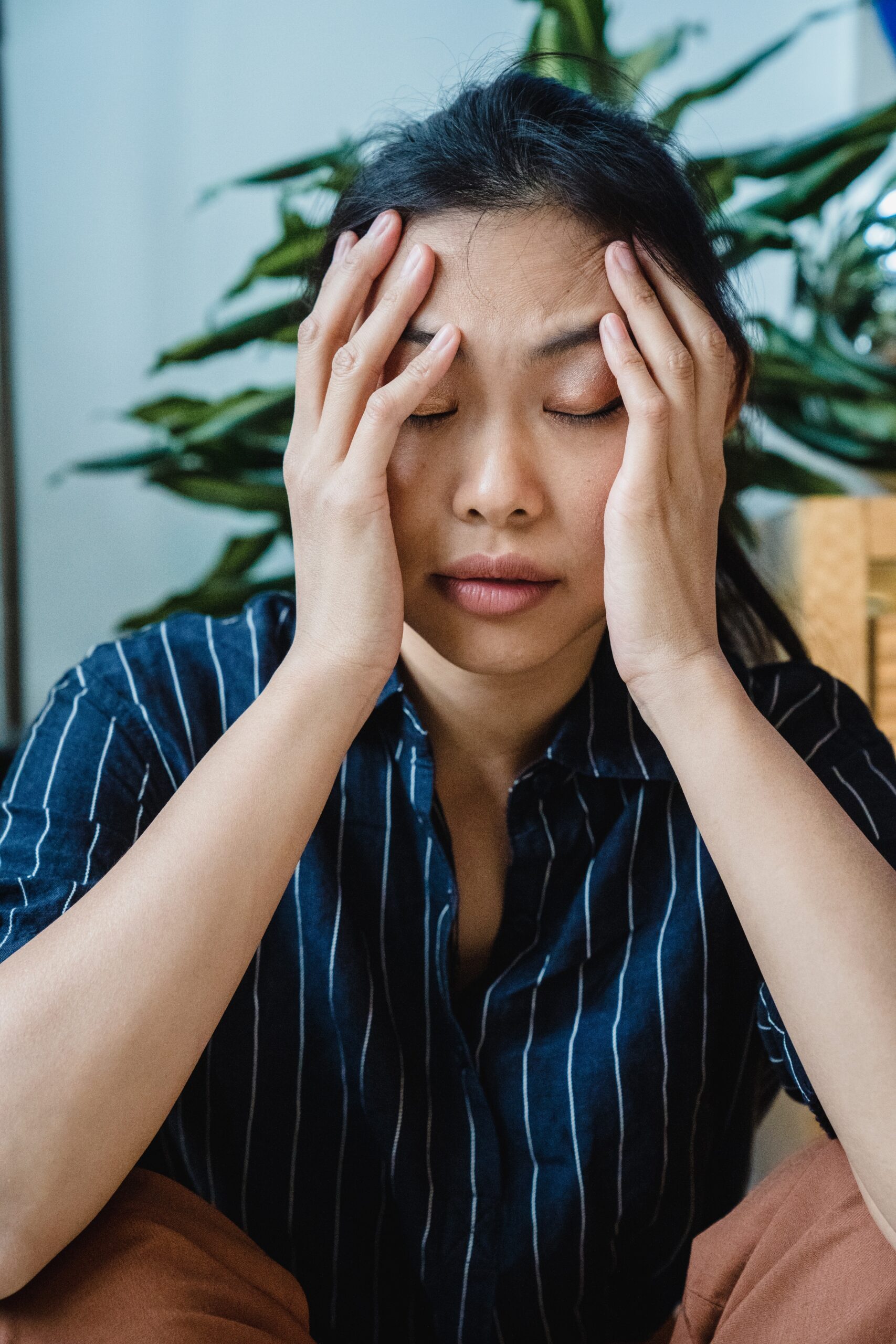 Woman with eyes closed, pressing temples, appears stressed; wearing blue striped shirt.