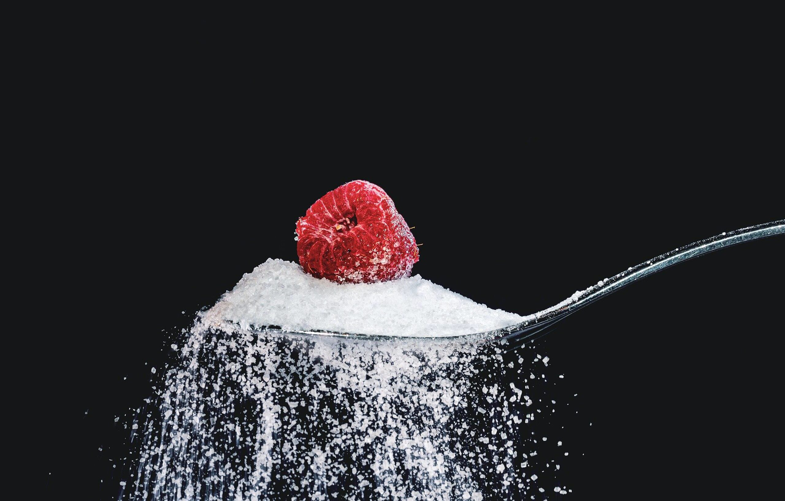 Red raspberry on a spoonful of sugar with sugar cascading against a black background