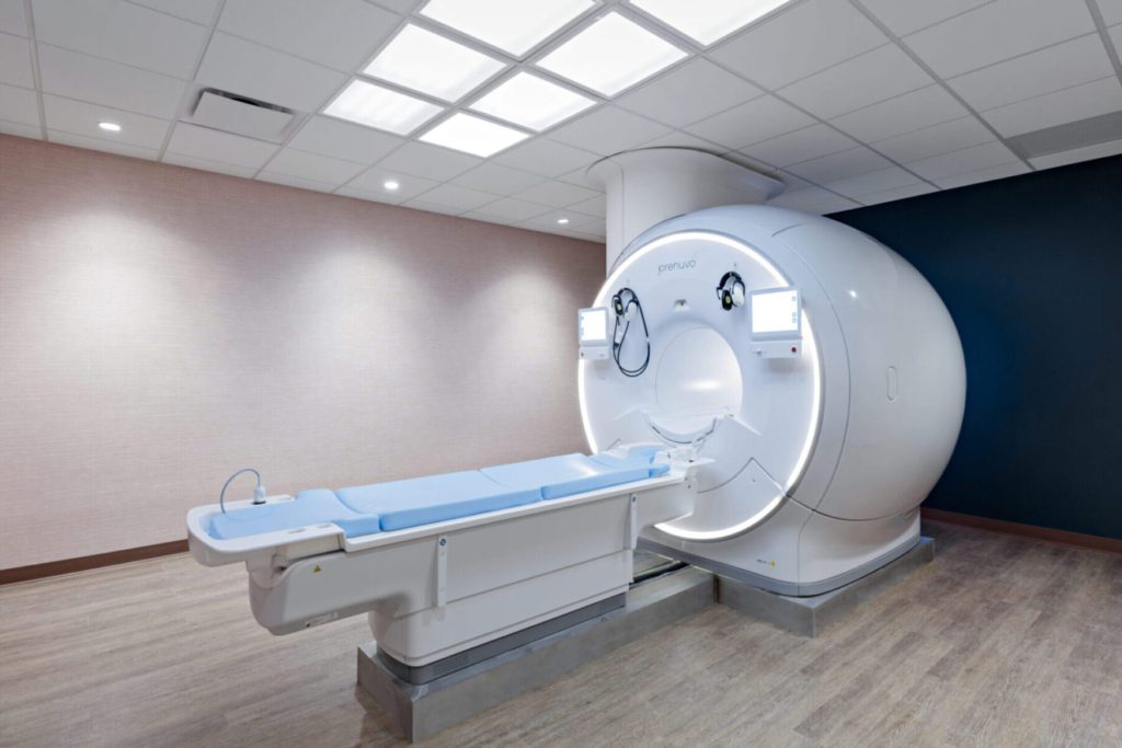 Large white MRI machine in a clinical room with an adjacent patient bed and soft lighting.