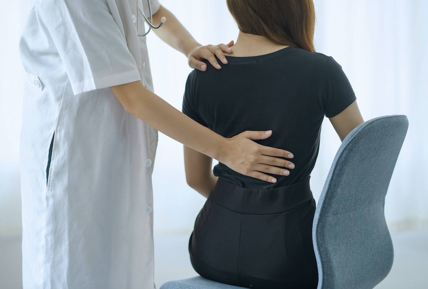 Women wearing a blue shirt sits with her back to the camera while a doctor examines her and places hand on her back.