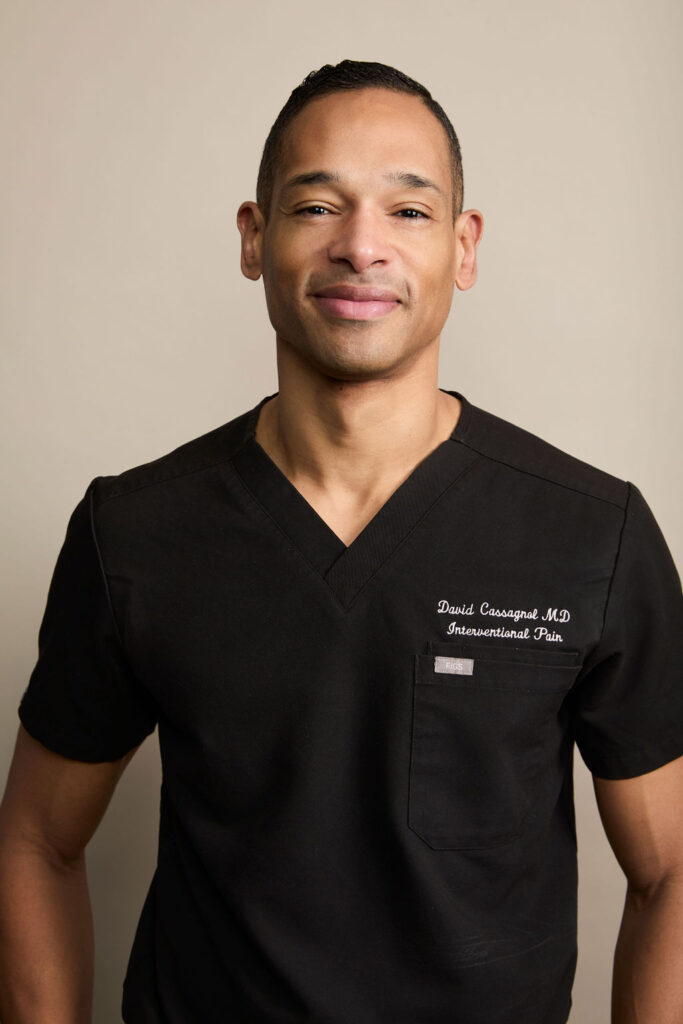 Man in black medical scrubs smiling softly against a beige background.