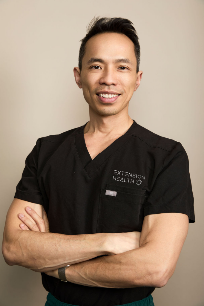 Smiling Asian man in black scrubs with “EXTENSION HEALTH” logo, arms crossed.