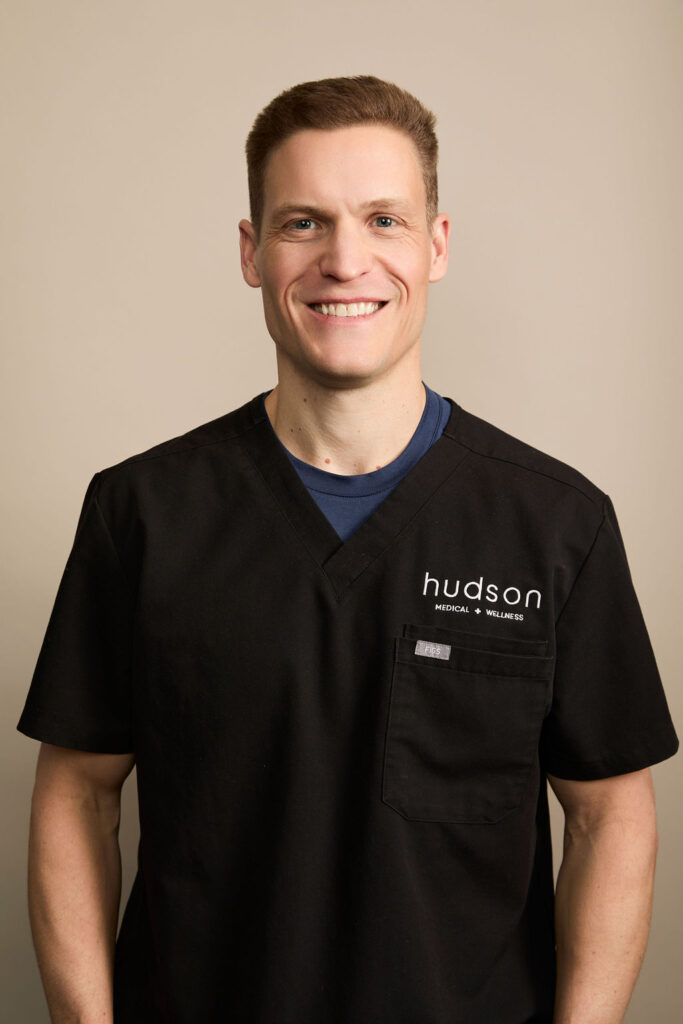 Smiling man in black medical scrubs with Hudson Medical Wellness logo on pocket, beige background.