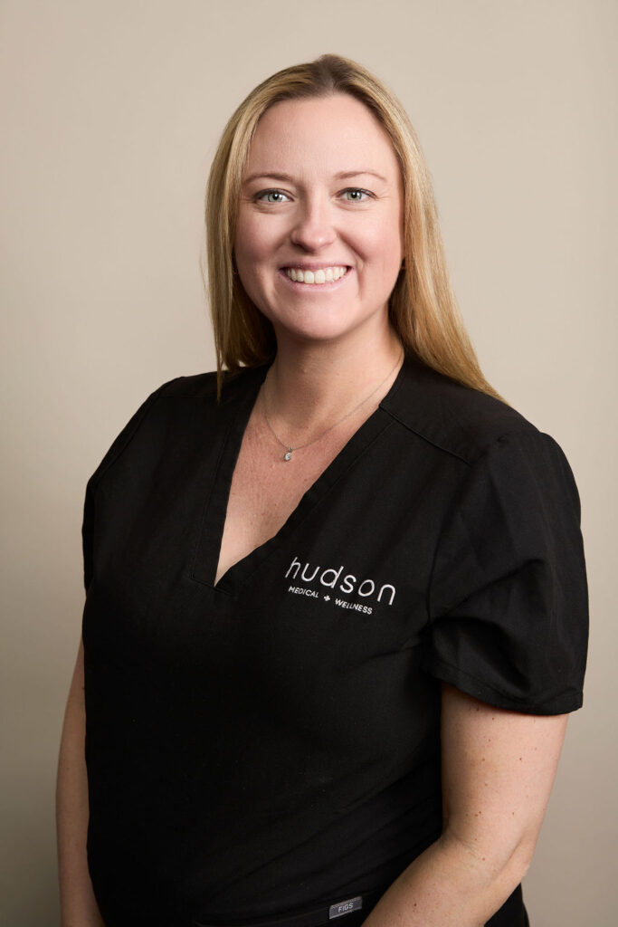 Smiling woman with long blonde hair in a black scrub top, beige background.