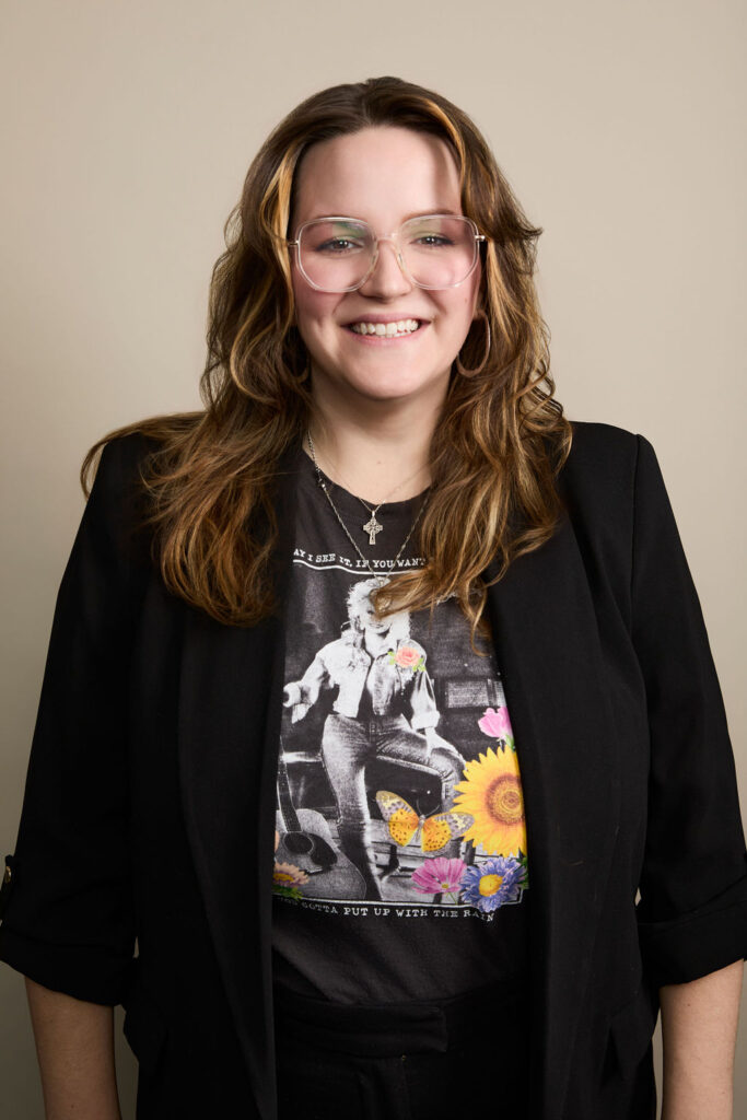 Smiling woman with glasses, wavy hair, black blazer, graphic t-shirt, beige background.