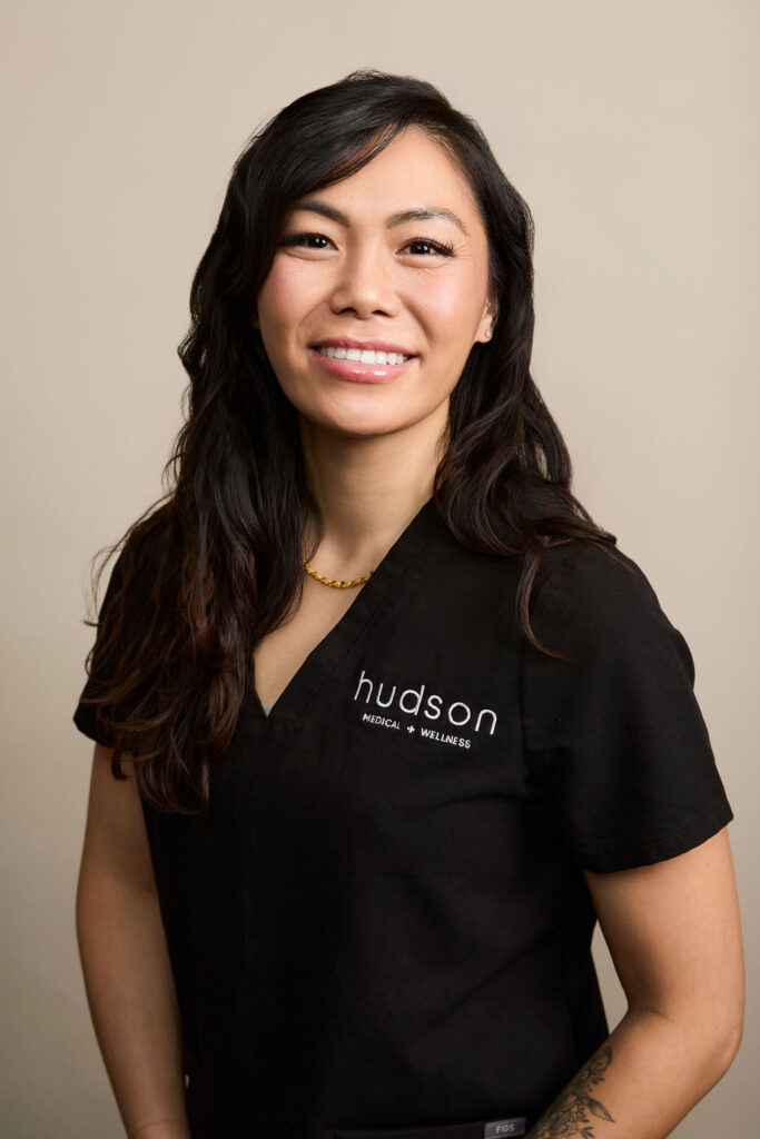 Portrait of smiling woman in black scrubs with Hudson Medical + Wellness logo, beige background.