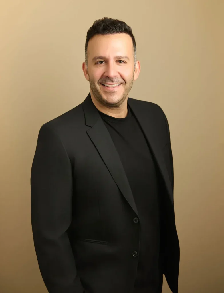 Smiling man in black blazer and shirt against beige background.